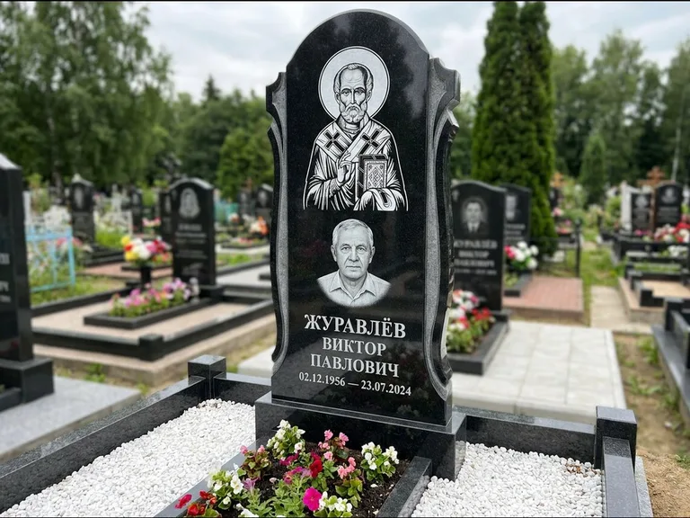 Памятник с гравировкой иконы: стела с образом в полный рост на обороте — гравировка образа Матроны Московской, низкие кусты спиреи в цвету, работа «Лебедева» в Норильске