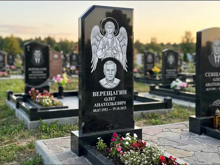 Памятник с гравировкой иконы: стела с резным навершием и нимбом в золоте — гравировка образа Спасителя, высокие разросшиеся туи полукругом, работа «Мельникова» в Норильске