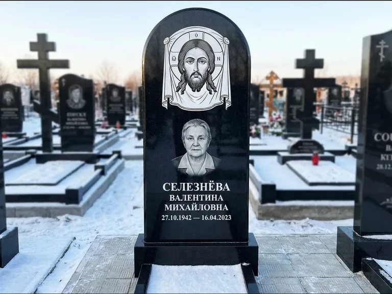 Памятник с гравировкой иконы: небольшая скромная одиночная стела с именной иконой — золочёный нимб на образе, аккуратный участок с газоном, работа «Ковалёв» в Норильске