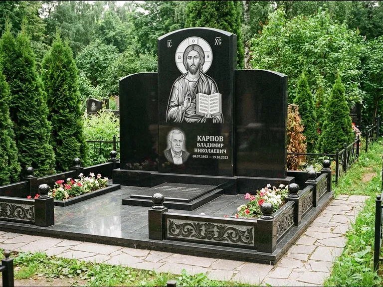 Памятник с гравировкой иконы: широкая панорамная стела с золочёным нимбом — гравировка образа Матроны Московской, цветник с белыми хризантемами, работа «Мельникова» в Норильске