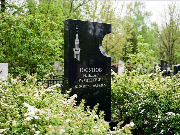 Мусульманский памятник с гравировкой мечети: стела с боковым вырезом в форме полумесяца — гравировка минарета с полумесяцем, низкие кусты спиреи в цвету, работа «Юсупов» в Норильске